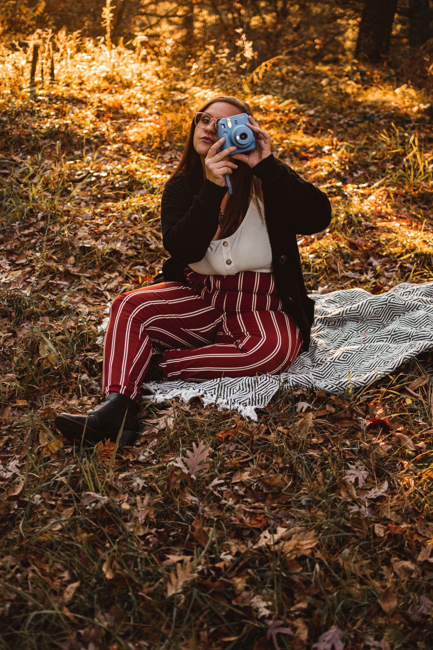 Person with camera sitting on blanket amid fall leaves during golden hour.