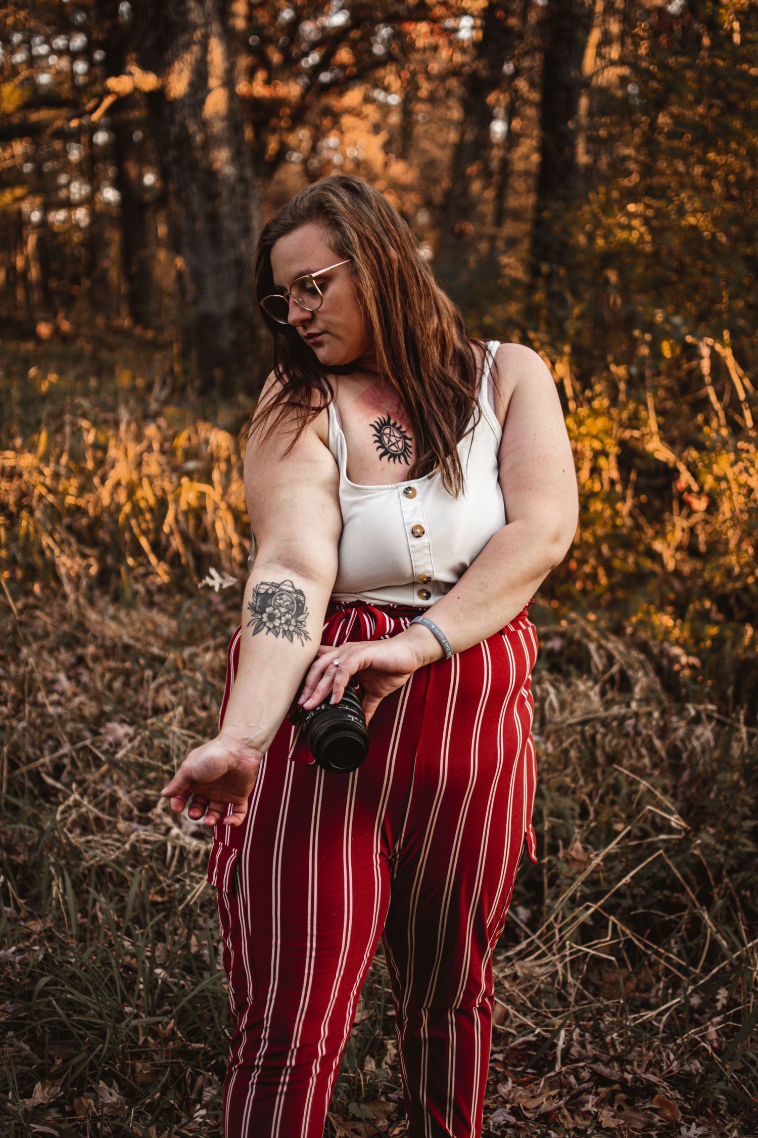Person in striped pants and white top holds camera in autumn forest.