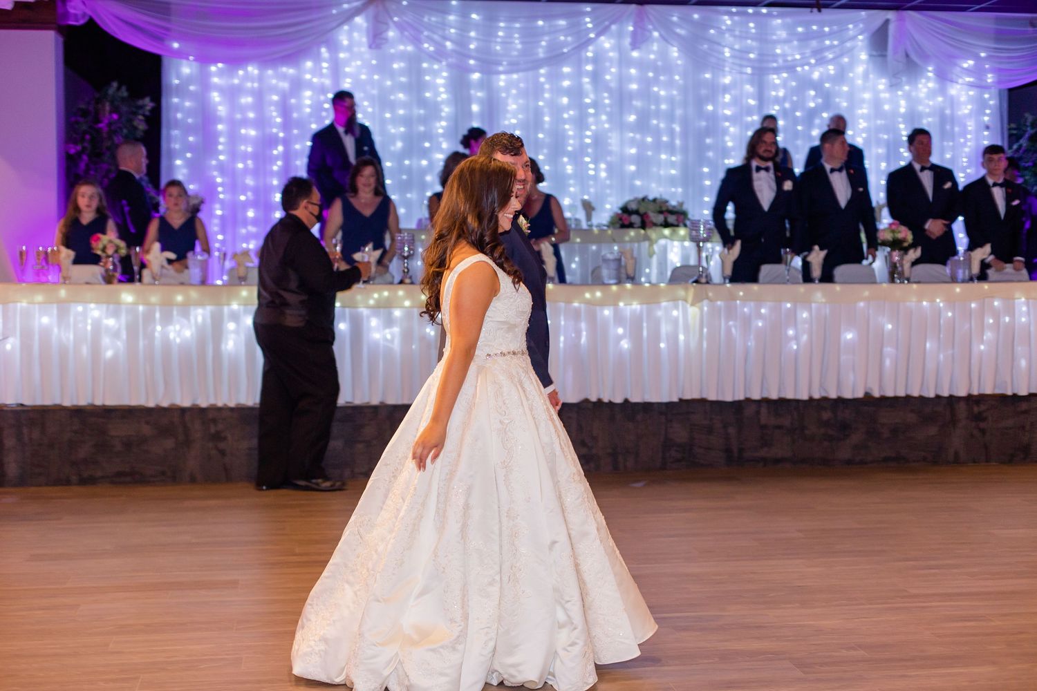 A bride in a white lace ballgown dances on a wooden floor with purple uplighting and twinkle lights in the background.