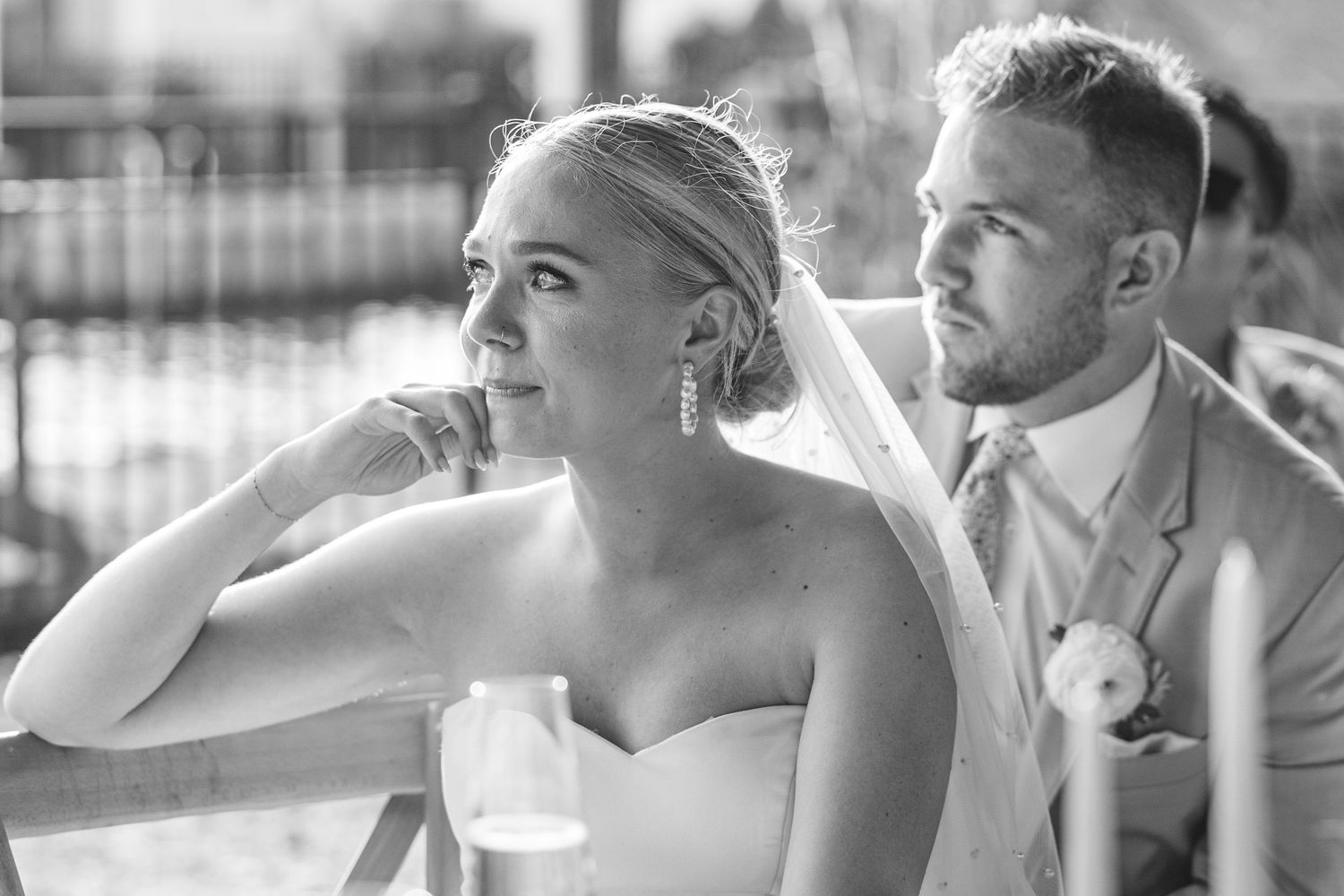 Bride wearing strapless dress touches earring while seated outdoors in black and white photo.