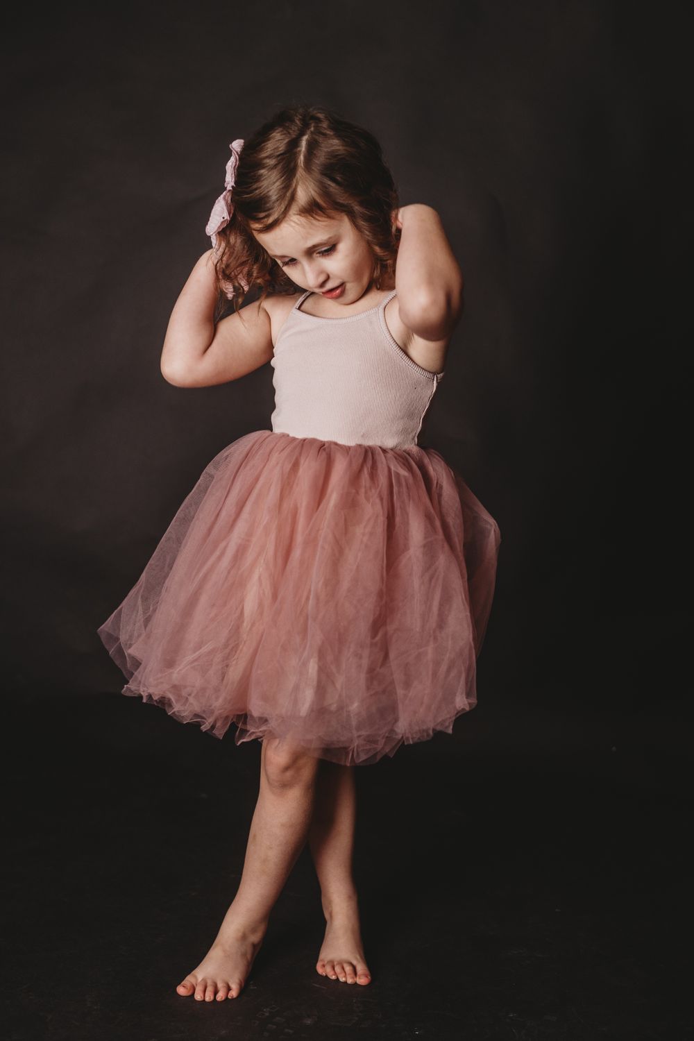 Child in pink tulle dress sitting and posing against dark backdrop.