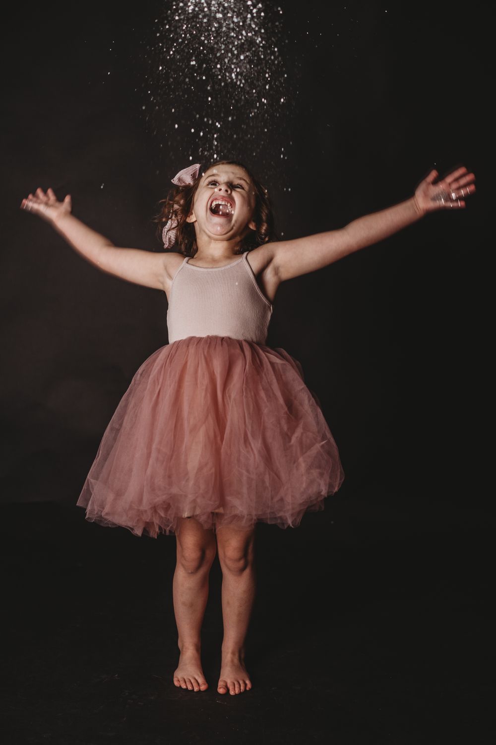 Child in pink tutu dress jumping and playing with glitter against black background.
