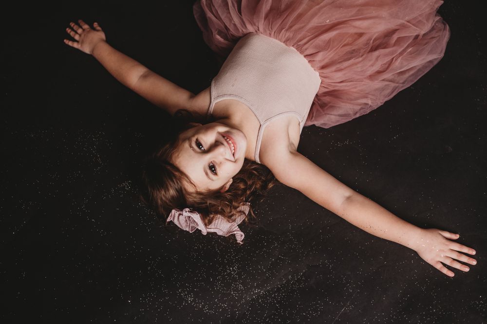 Child in pink dress lying on dark floor during artistic photo session.