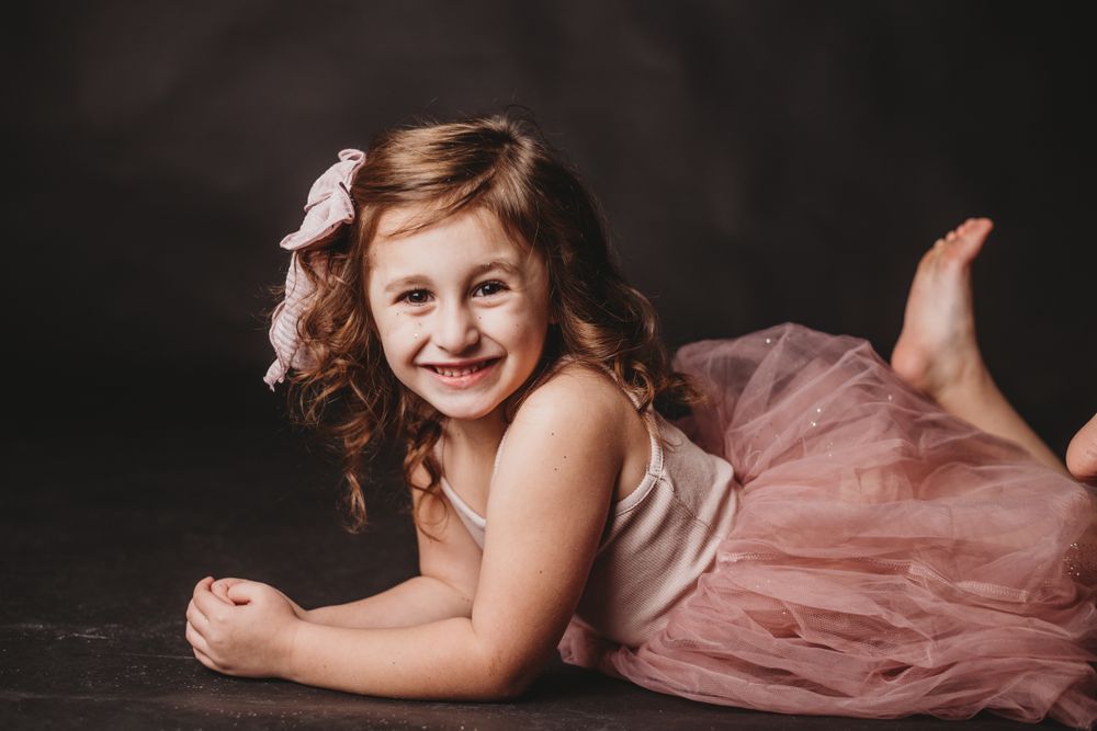 Studio portrait featuring child in pink tulle dress on dark background.