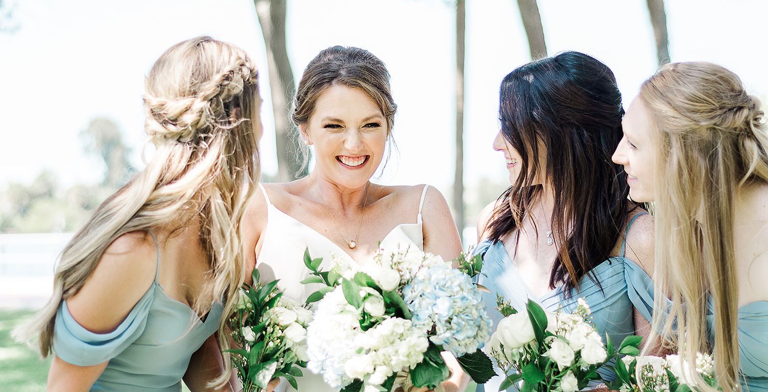 Bride and bridesmaids in light blue dresses share laughter while holding bouquets.