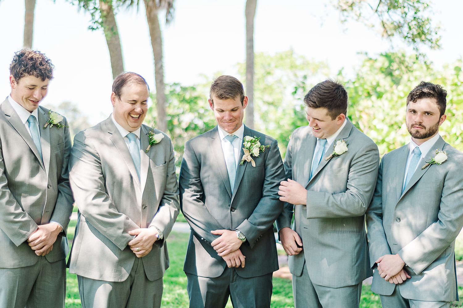 Groomsmen in light gray suits share candid moment during outdoor wedding.