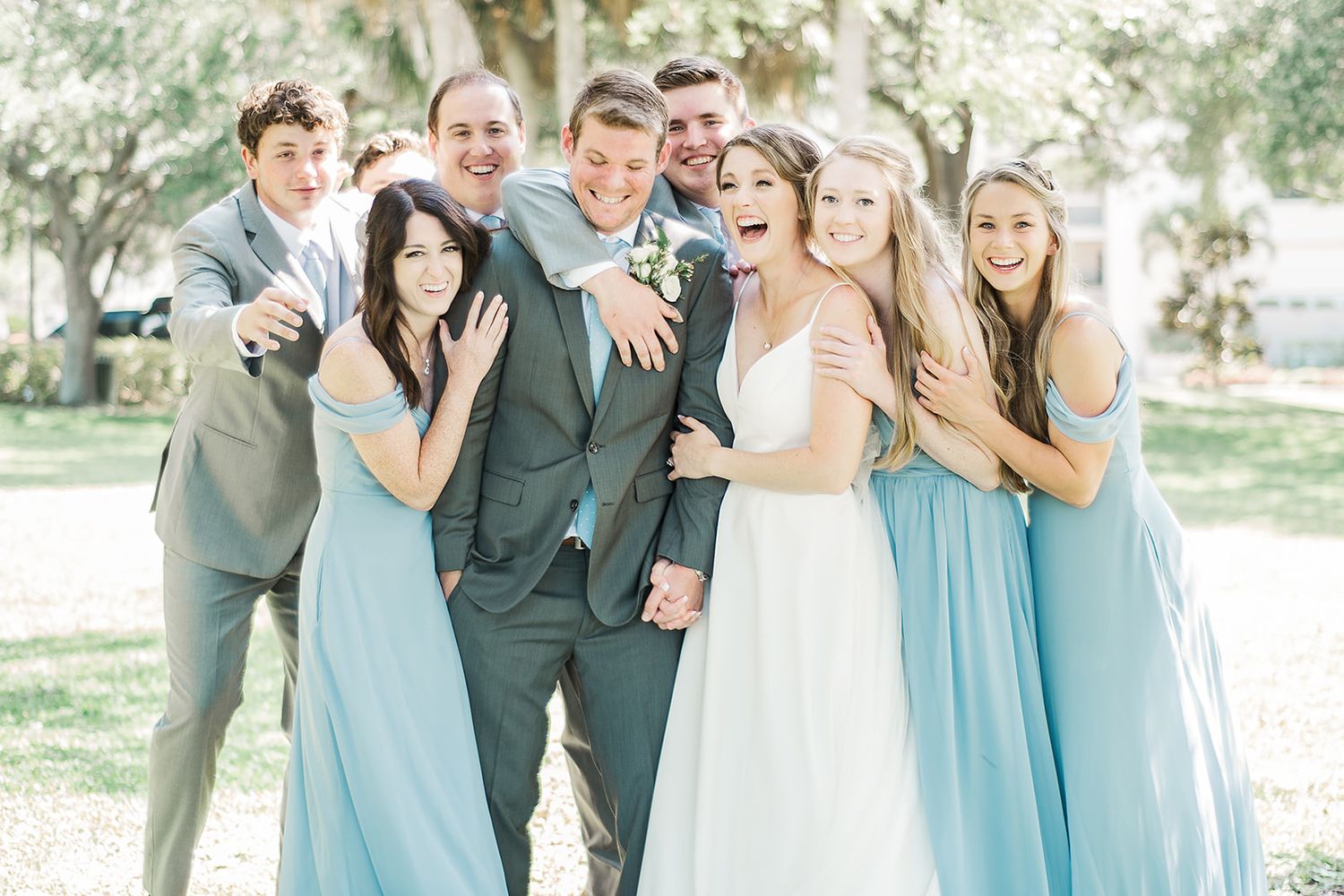 Wedding party in light blue bridesmaid dresses and gray suits pose playfully together outdoors.