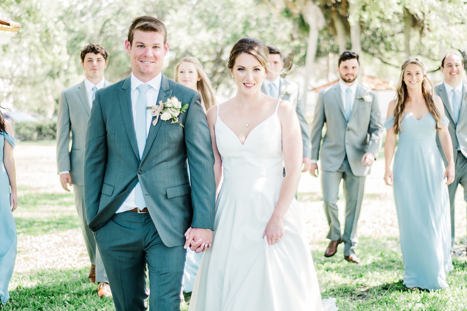 Wedding couple leads bridal party wearing light blue dresses and gray suits in outdoor garden setting.