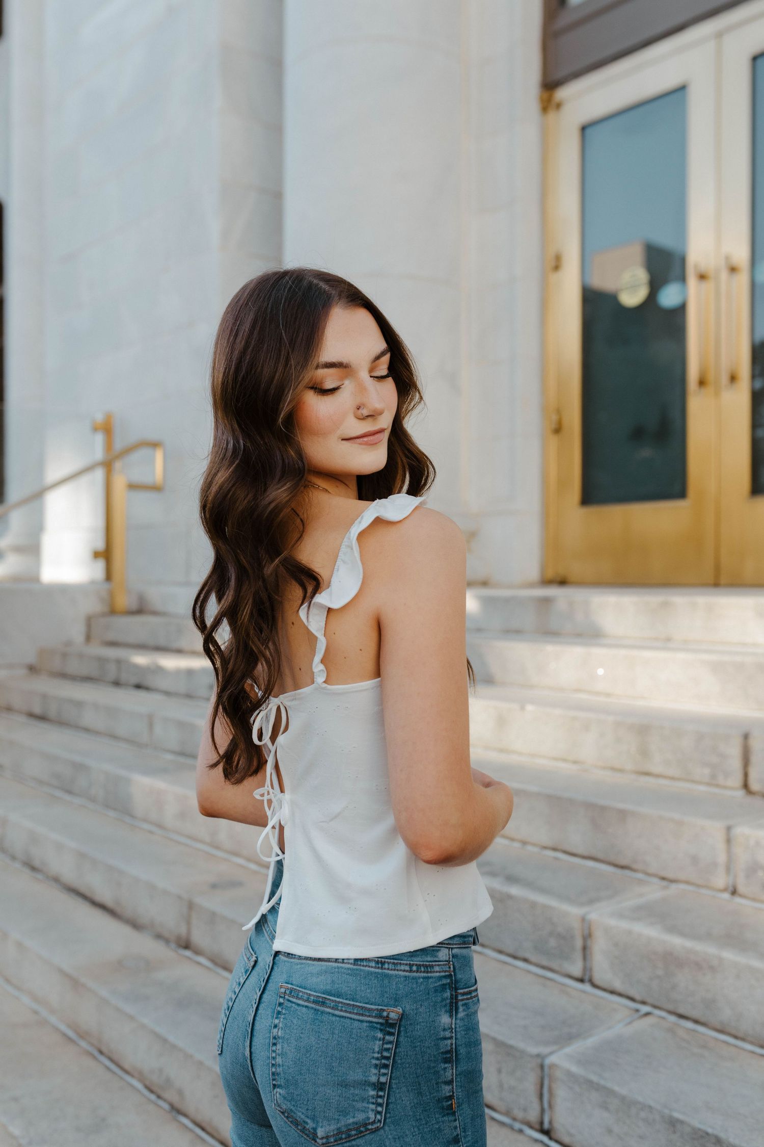 Woman in white ruffled top and jeans poses on stone steps in front of golden doors.