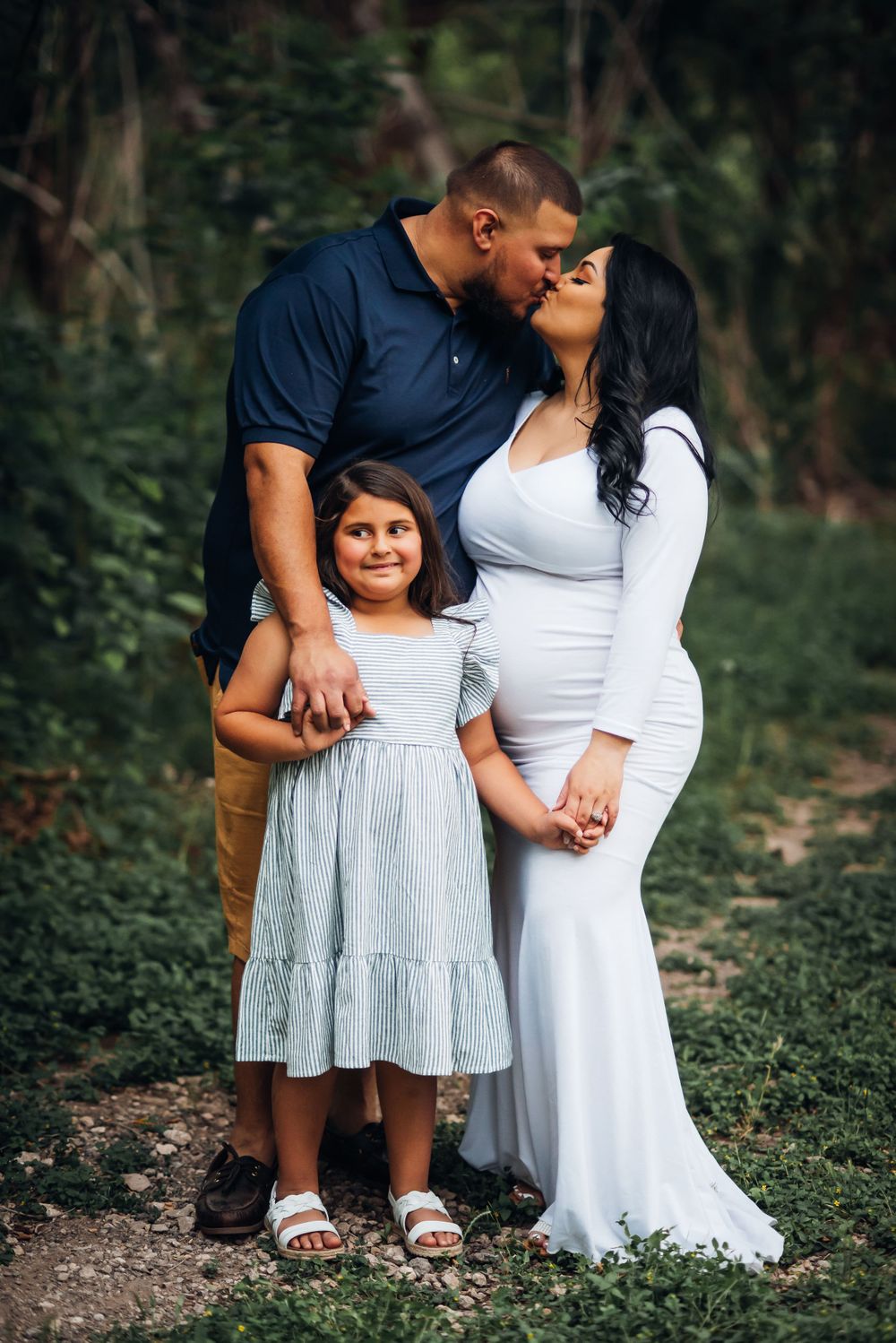 A family maternity portrait in a wooded setting with coordinated white and blue outfits.