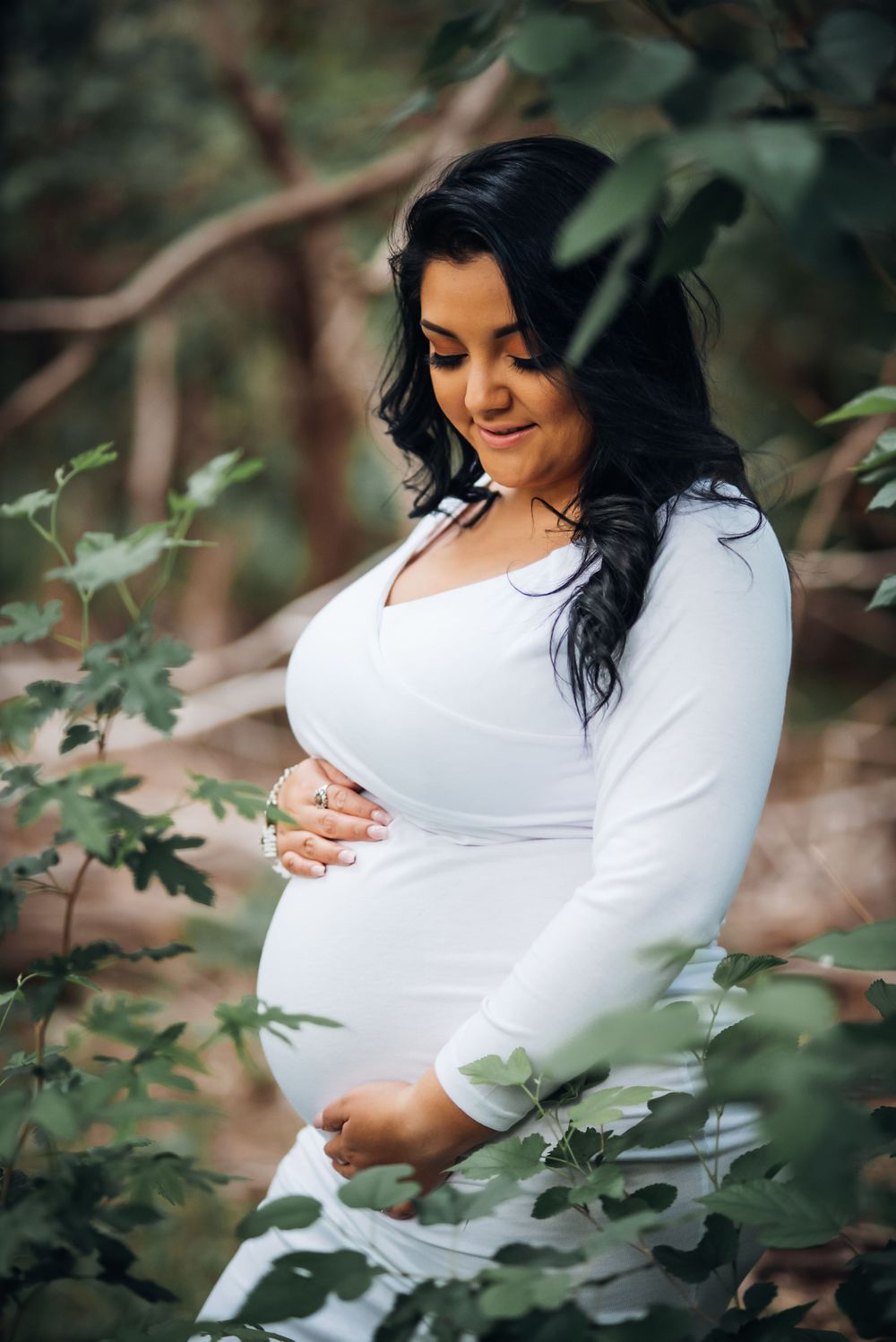 An expectant mother in a white dress poses thoughtfully among leafy branches.