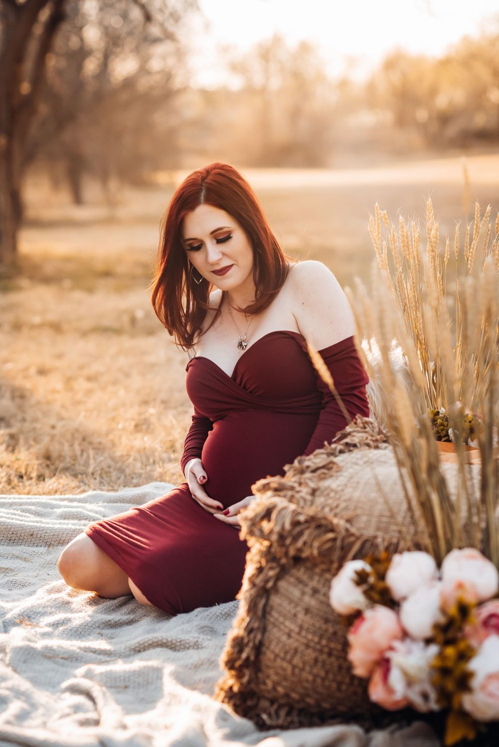 A maternity photo featuring a burgundy dress against warm sunset lighting with dried pampas grass.