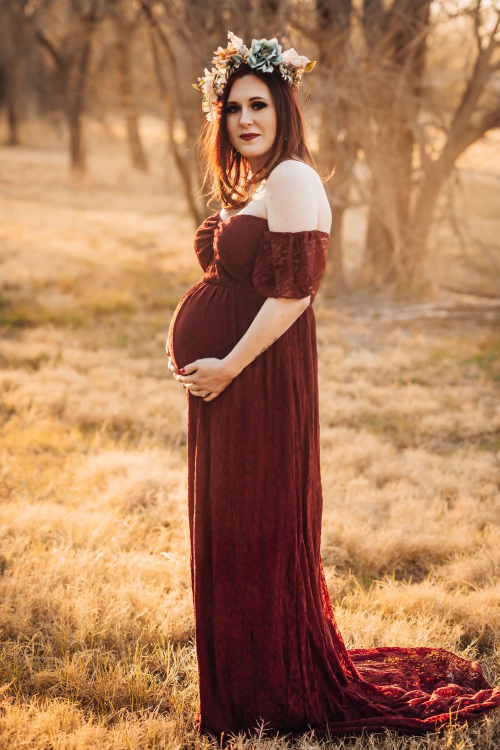 A romantic maternity portrait in a flowing burgundy dress with floral crown at golden hour.