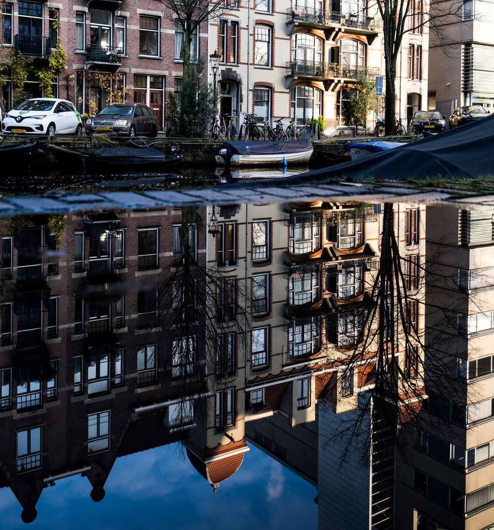 Amsterdam canal reflecting tall brick residential buildings and bare trees on a sunny day.