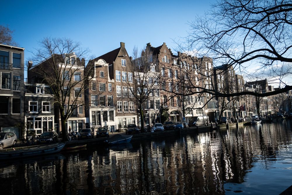 Classic Amsterdam canal houses with brick facades reflecting in calm water on a sunny winter day.