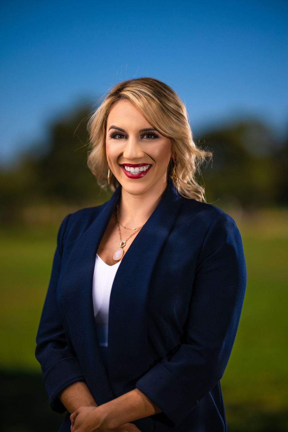 Professional headshot of woman in navy blazer and white top against outdoor background.