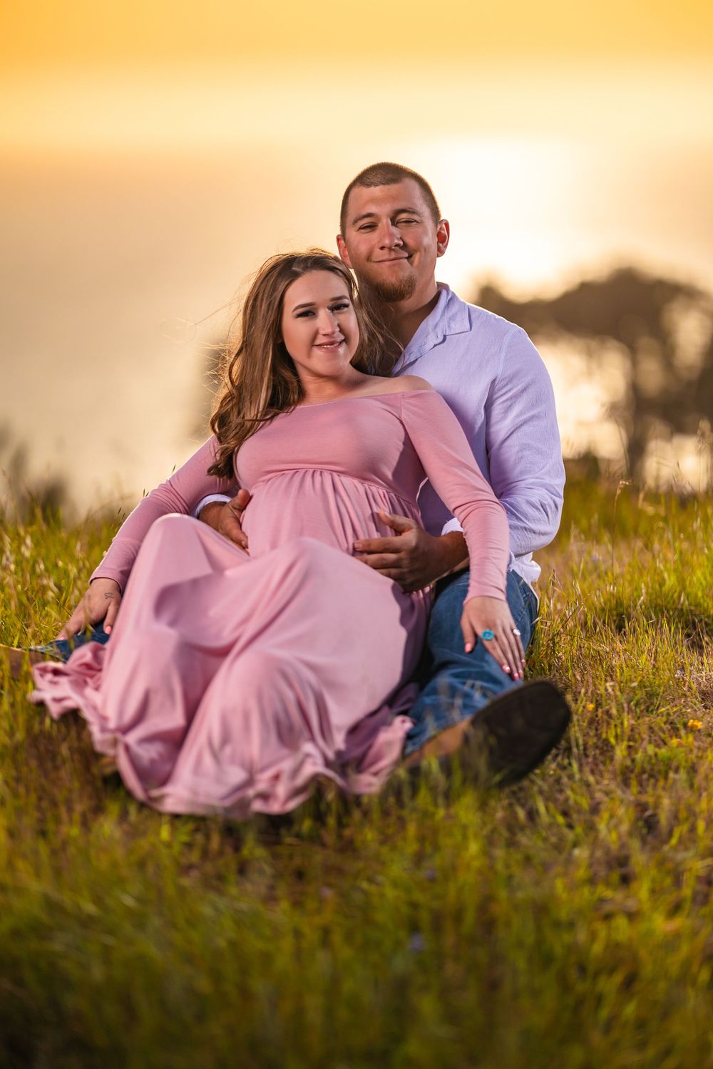 A couple in casual attire sits together in a grassy field during golden hour sunset.
