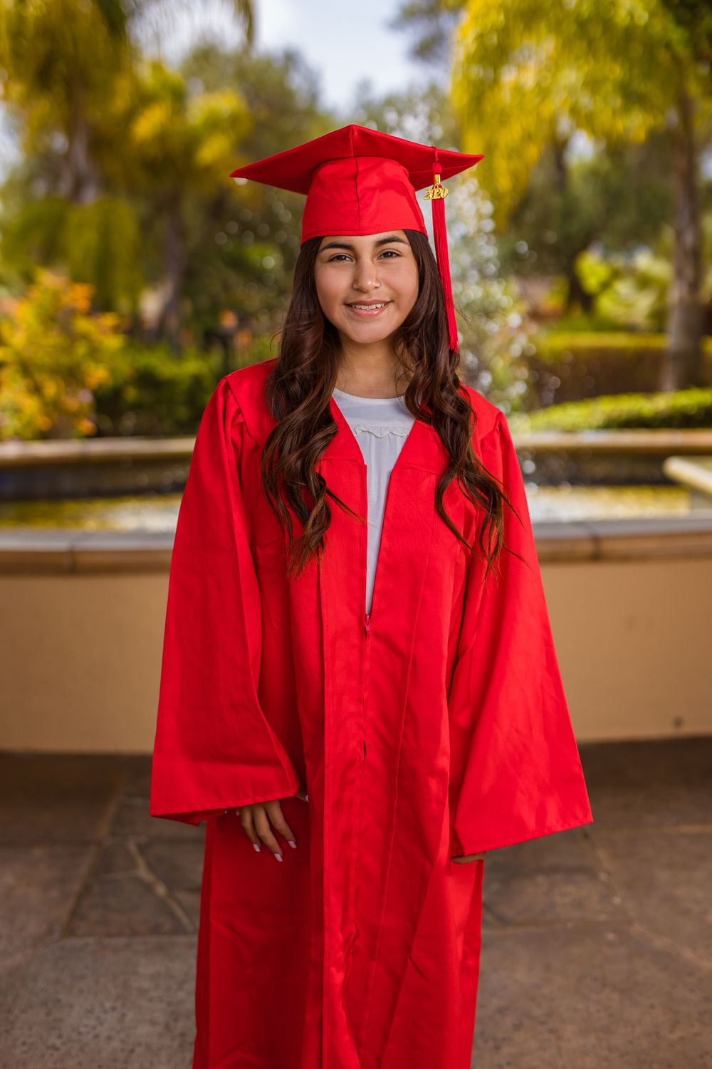 A graduate poses in traditional bright red cap and gown against a garden backdrop with fountains.