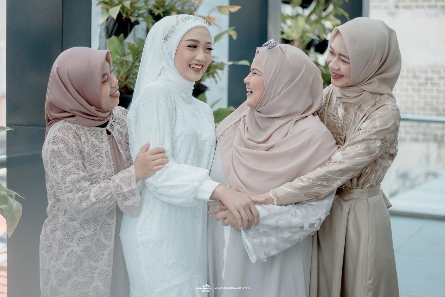 Four women in pastel hijabs and formal dresses sharing a joyful moment together outdoors.