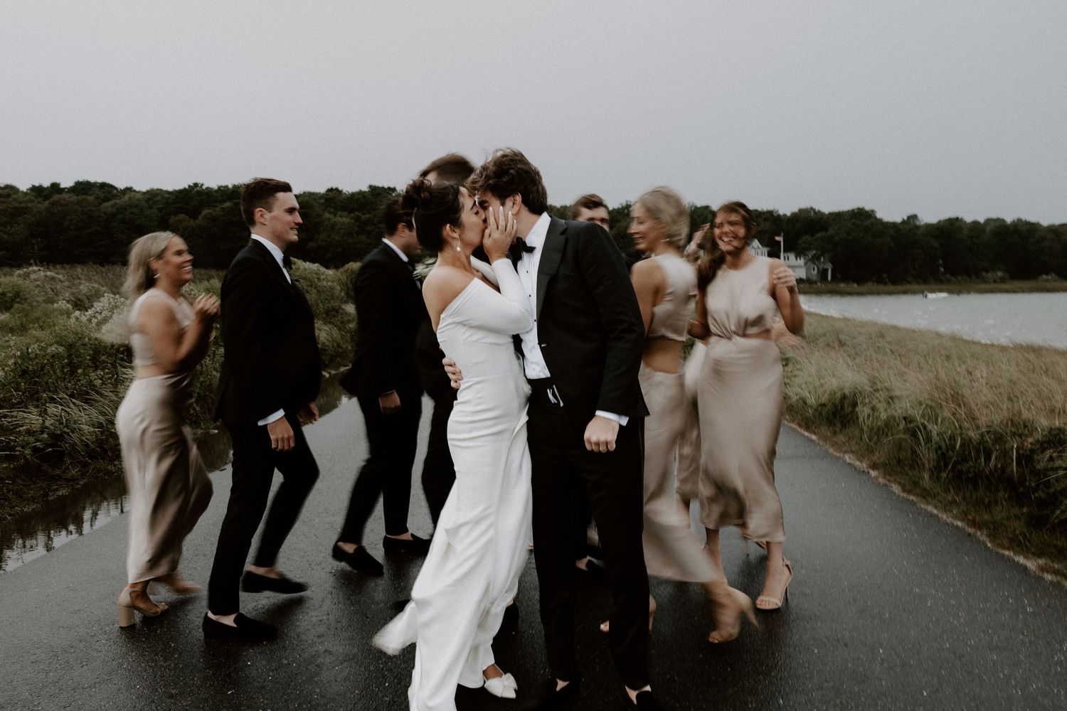 Wedding party walking together on a rainy road, bride and groom kissing surrounded by guests. Cape Cod, Ma. Miranda Photo & Co