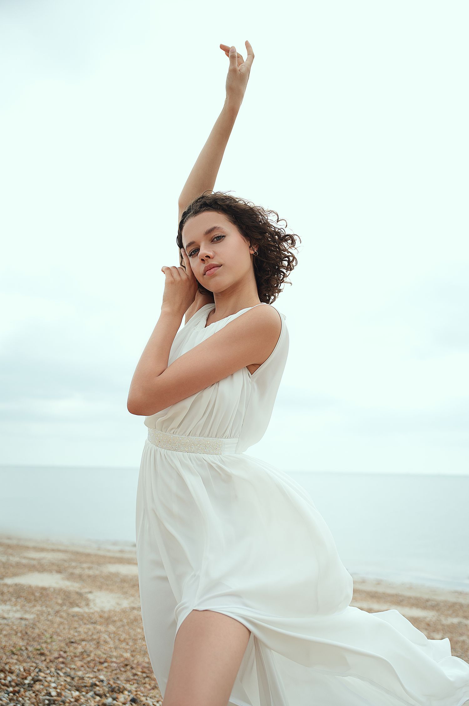 Dancing figure in flowing white dress against cloudy coastal backdrop.