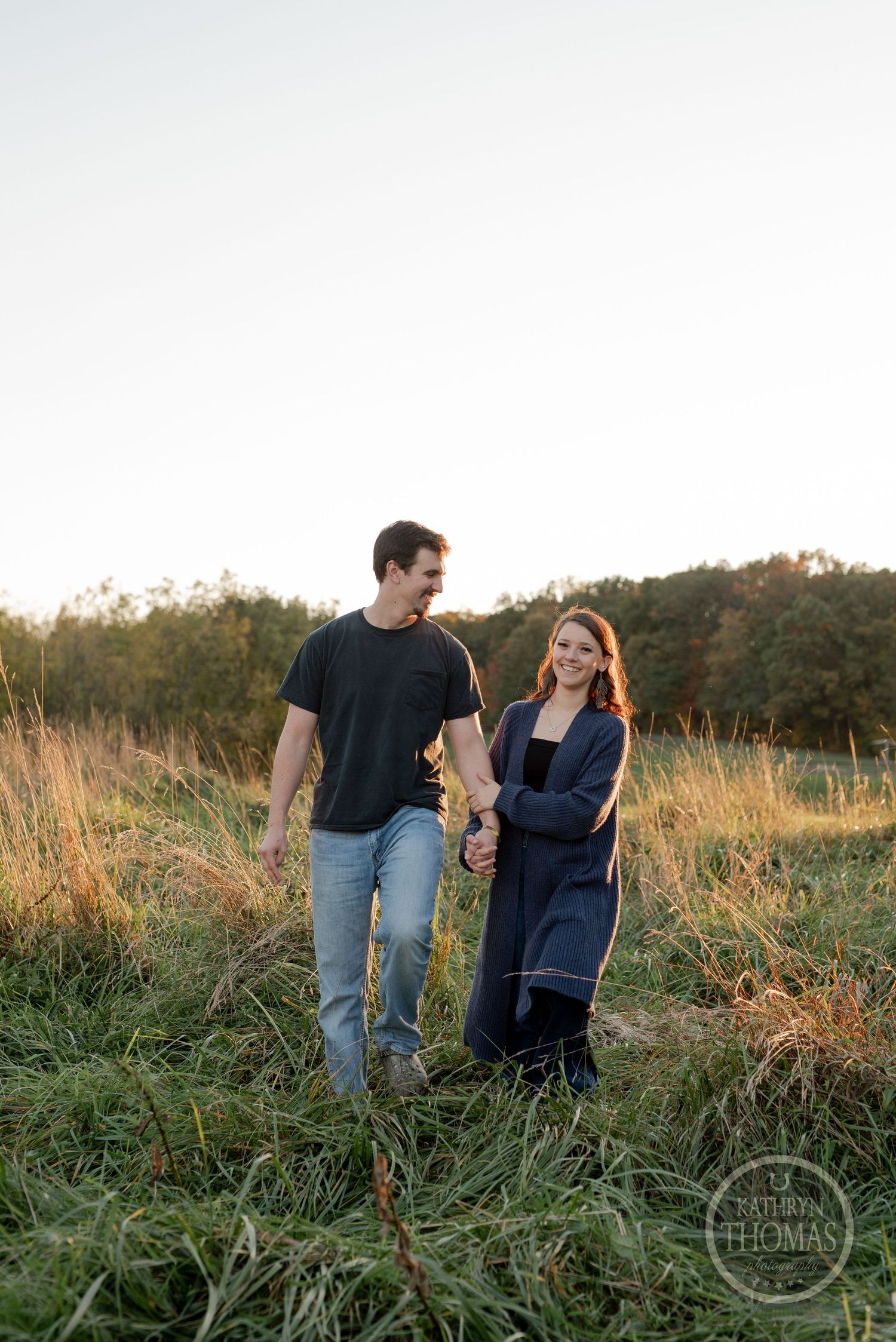 A couple walks hand in hand through tall grass during golden hour.