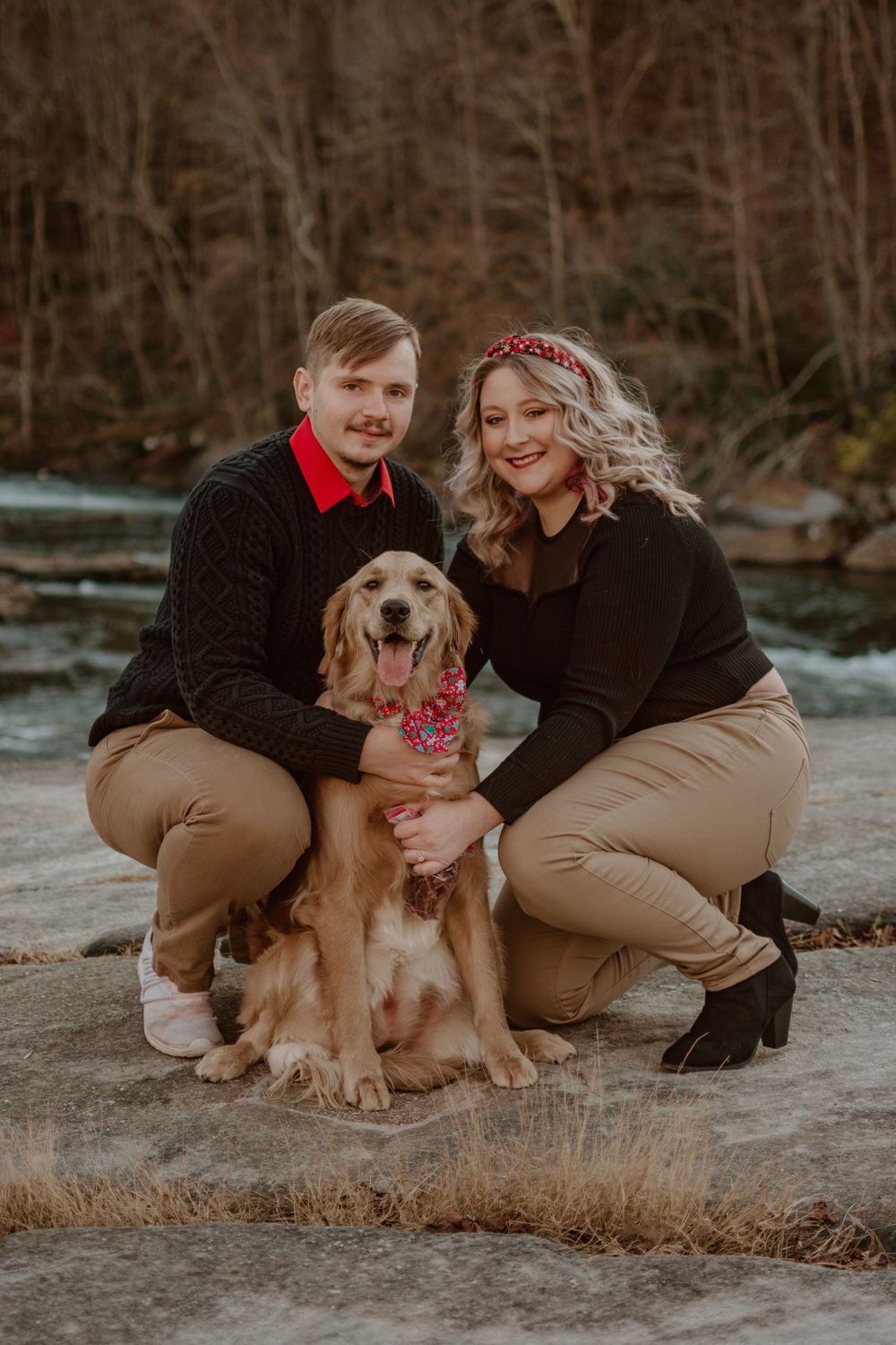 A couple poses with their golden retriever wearing a festive bow on a winter day.