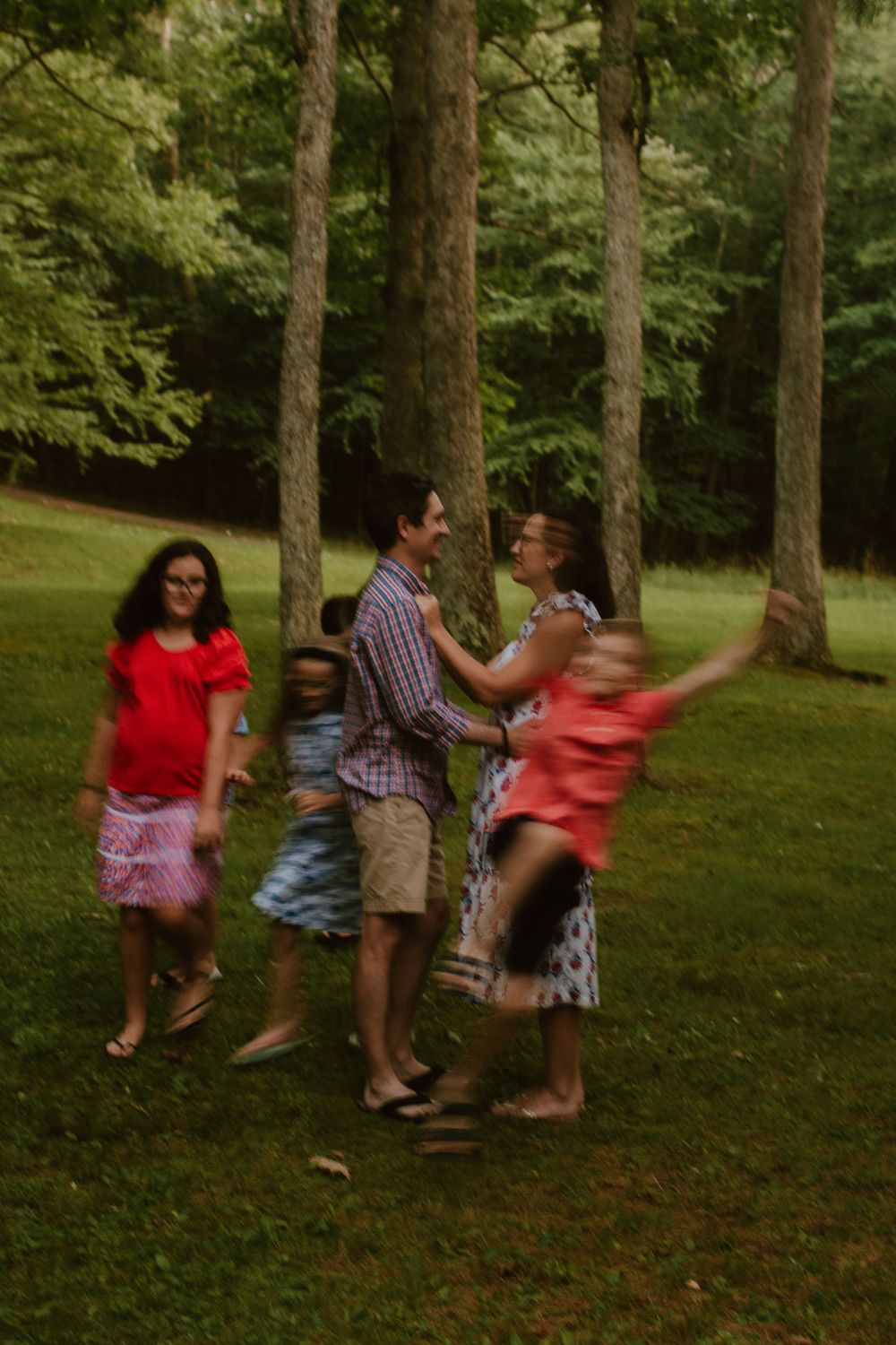 Family playing and having fun together under tall trees in a grassy park setting.
