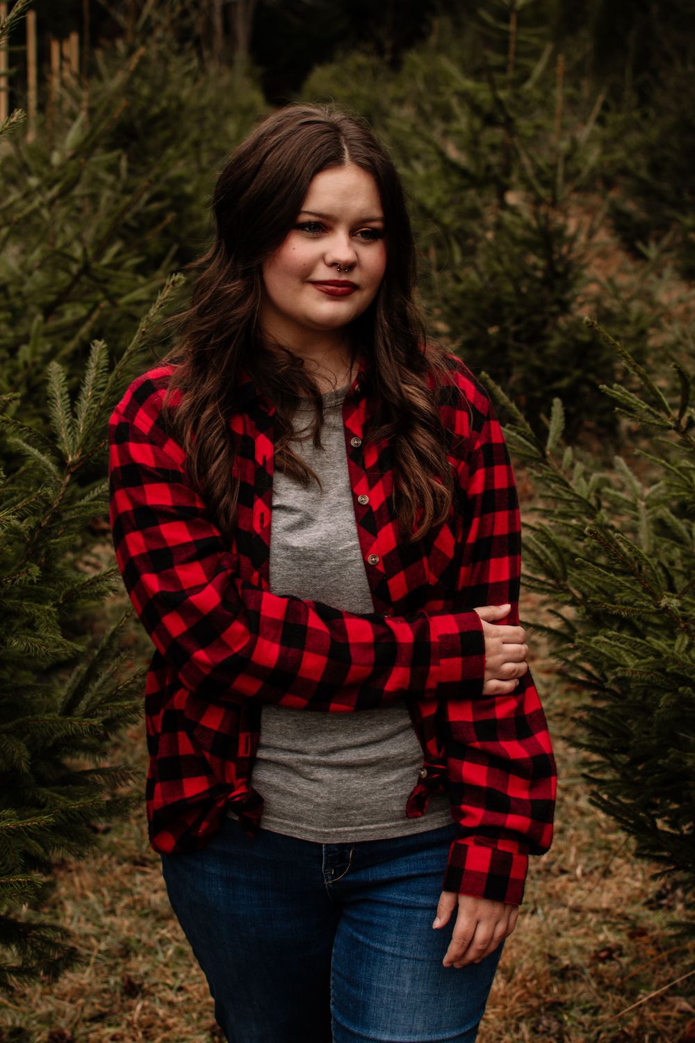 Person in red buffalo plaid shirt and jeans poses casually at a Christmas tree farm.