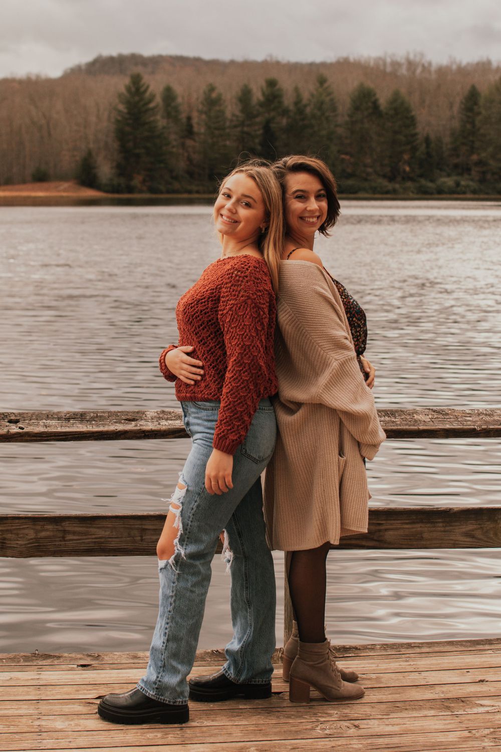 Two people posing playfully back-to-back on a wooden dock with a lake in the background.