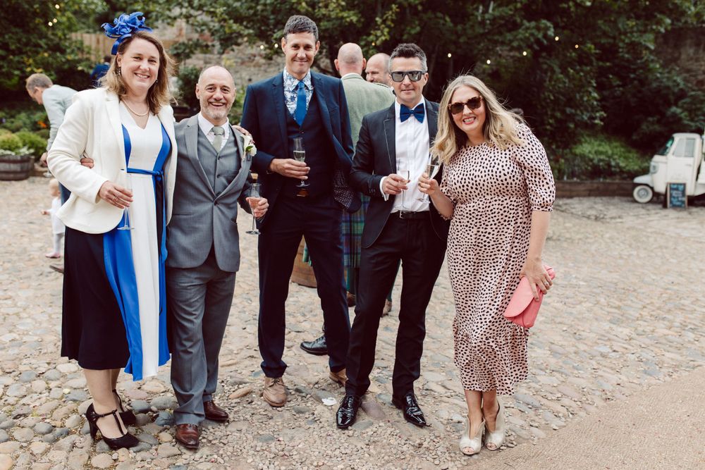 Wedding guests in formal attire gathered outside a rustic stone building.