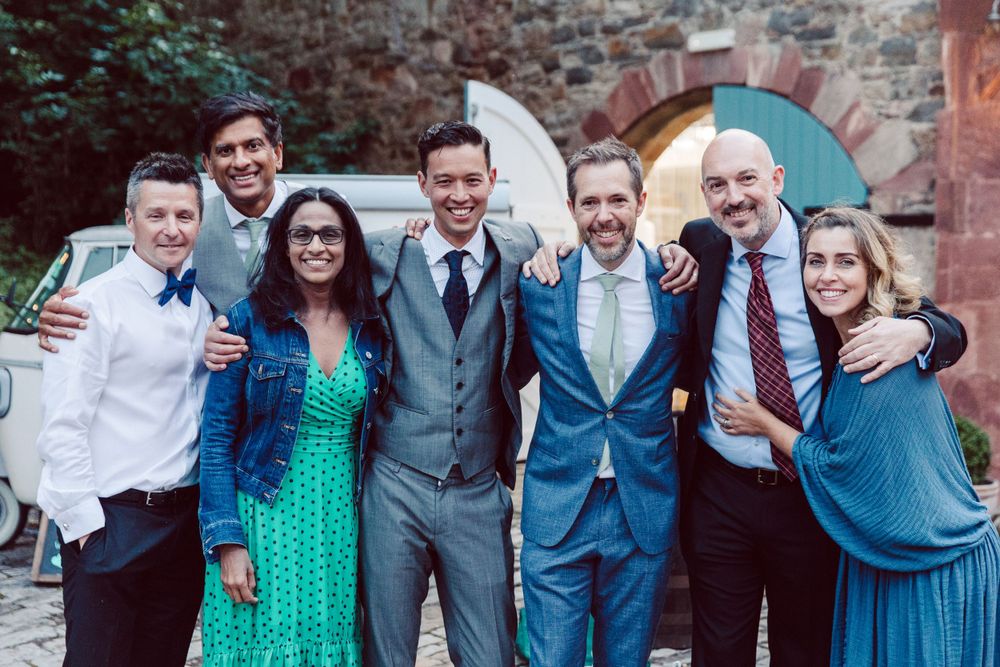 Group photo of wedding attendees in blue and green attire laughing together outdoors.