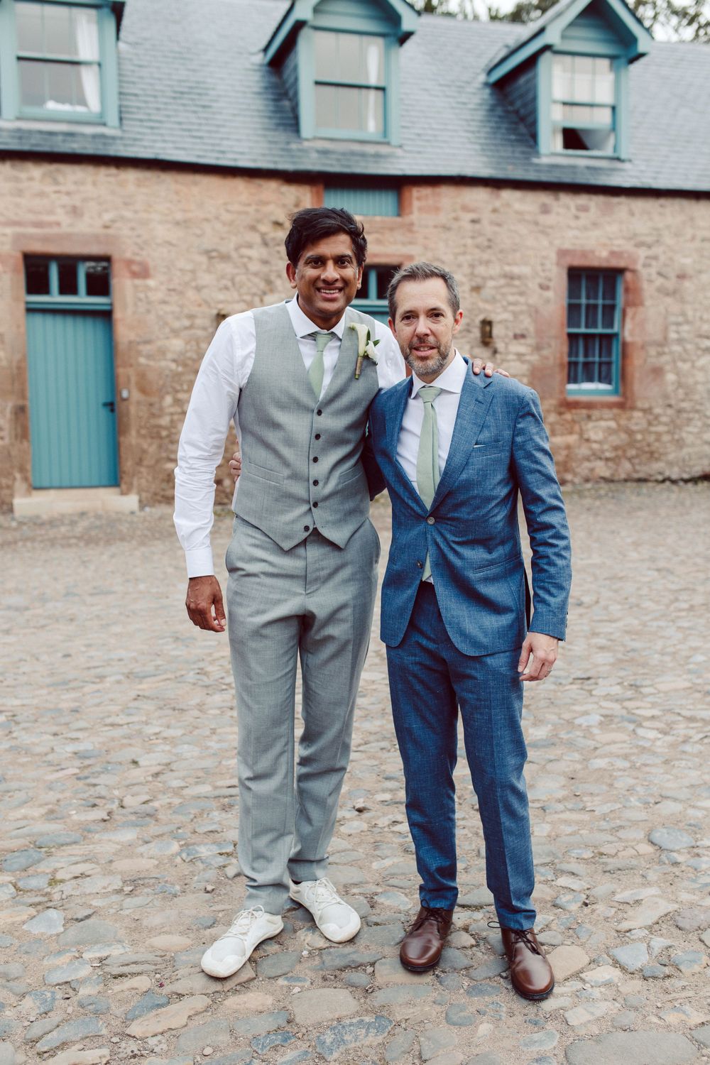 Groomsmen in suits posing casually against historic stone building exterior.