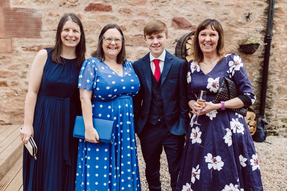 Group photo of wedding guests wearing coordinated blue and navy dresses against stone wall.