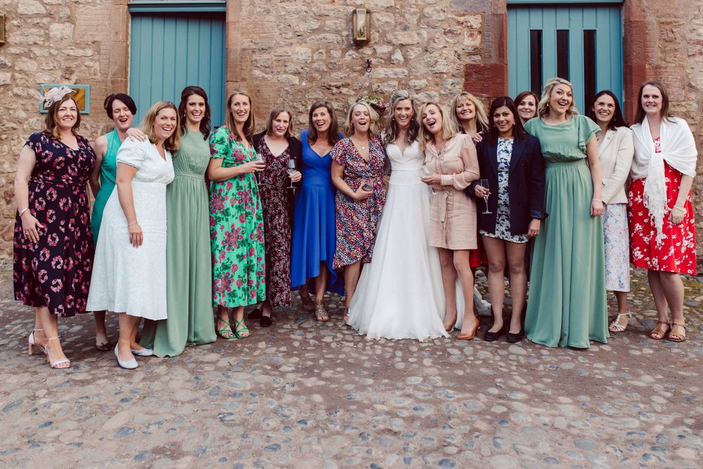 Large wedding group photo with guests lined up against rustic stone building facade.