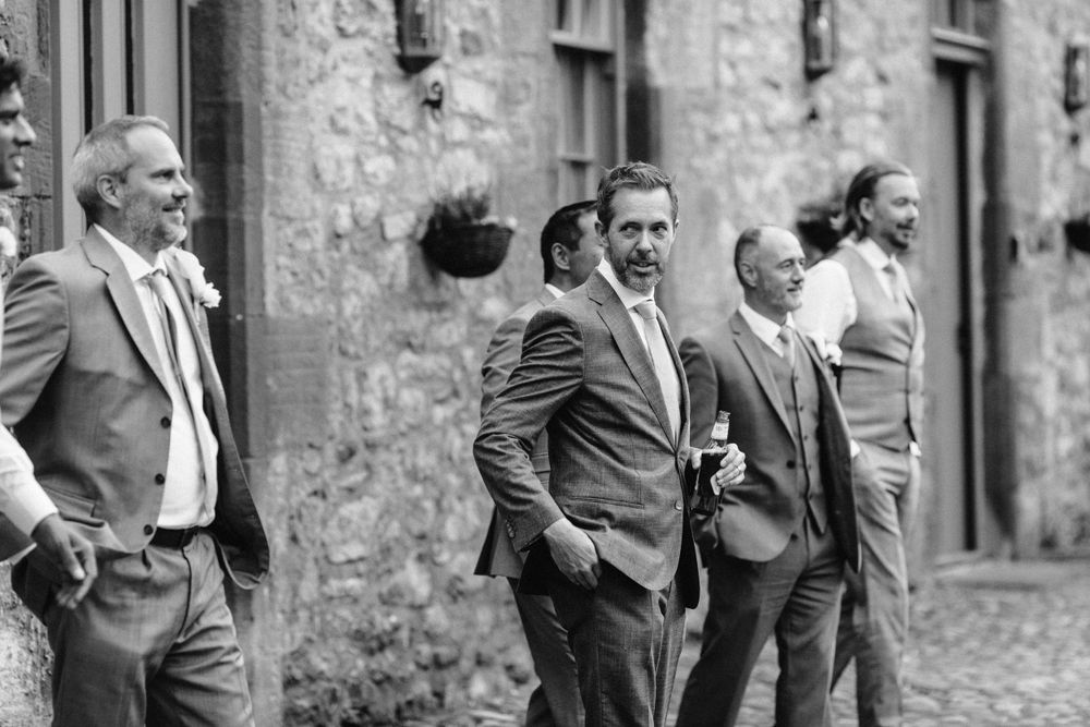 Black and white photo of wedding guests mingling outside historic stone building.