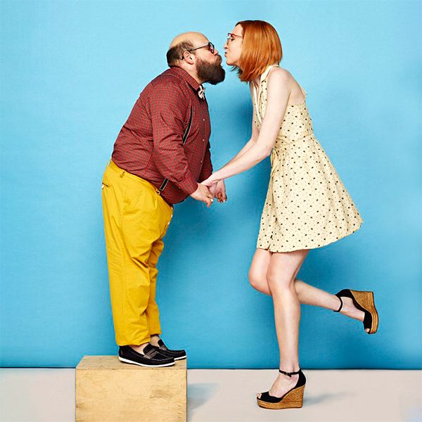 Playful couple posing for quirky kiss on wooden block against bright blue backdrop.