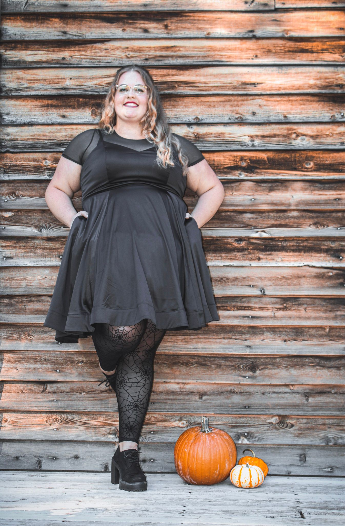 Autumn portrait with black dress and decorative pumpkins against weathered wooden wall.