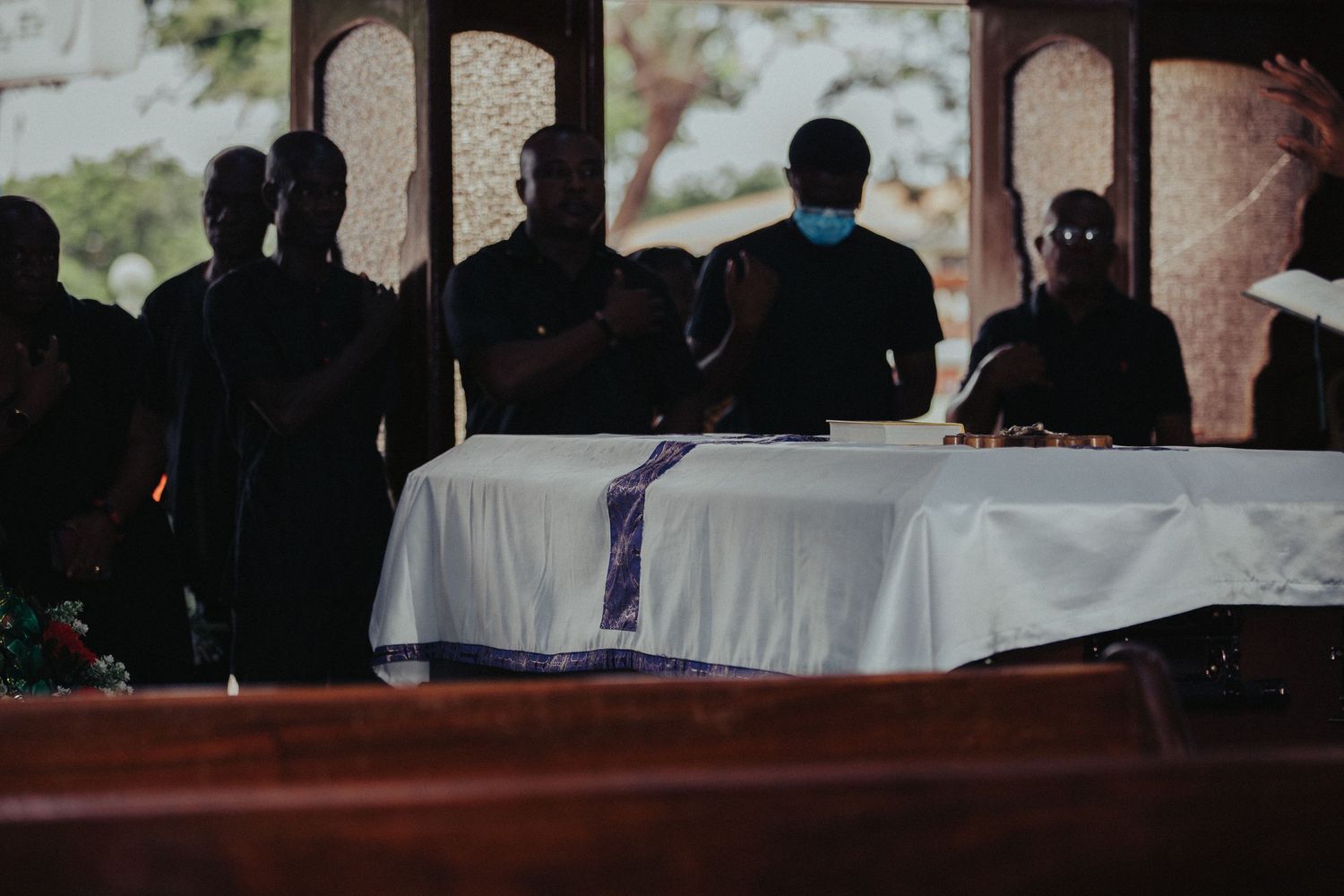 Group of mourners gathered around a white casket during a solemn indoor funeral ceremony.