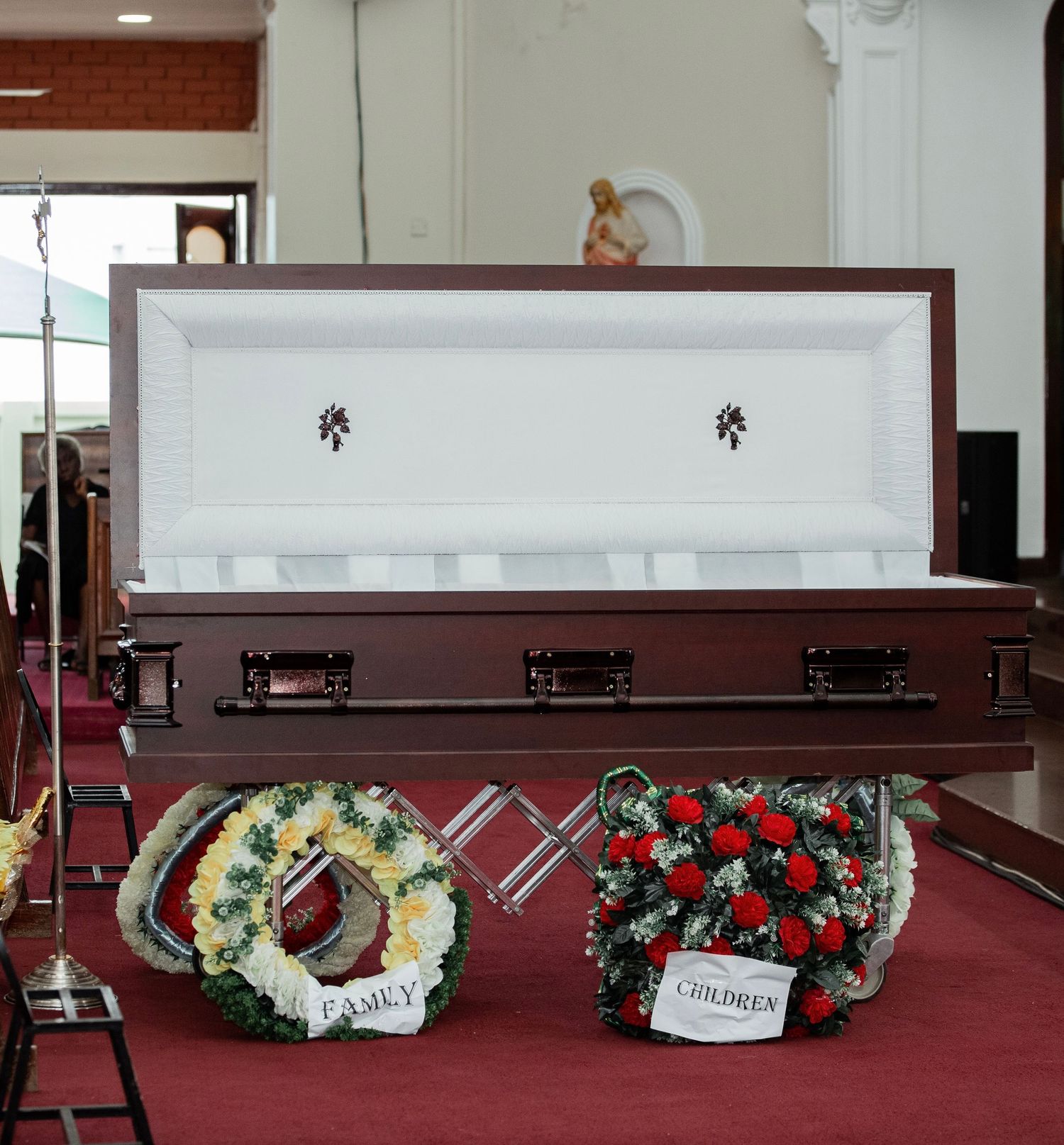 View of a casket at the front of a church altar surrounded by white floral wreaths and a memorial cross.