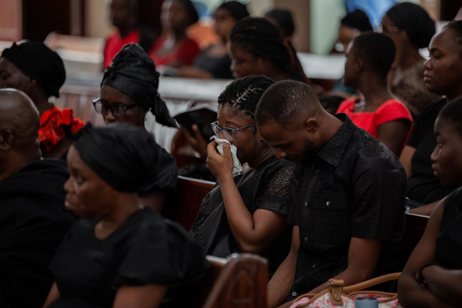 Group of mourners gathered around a white casket during a solemn indoor funeral ceremony.