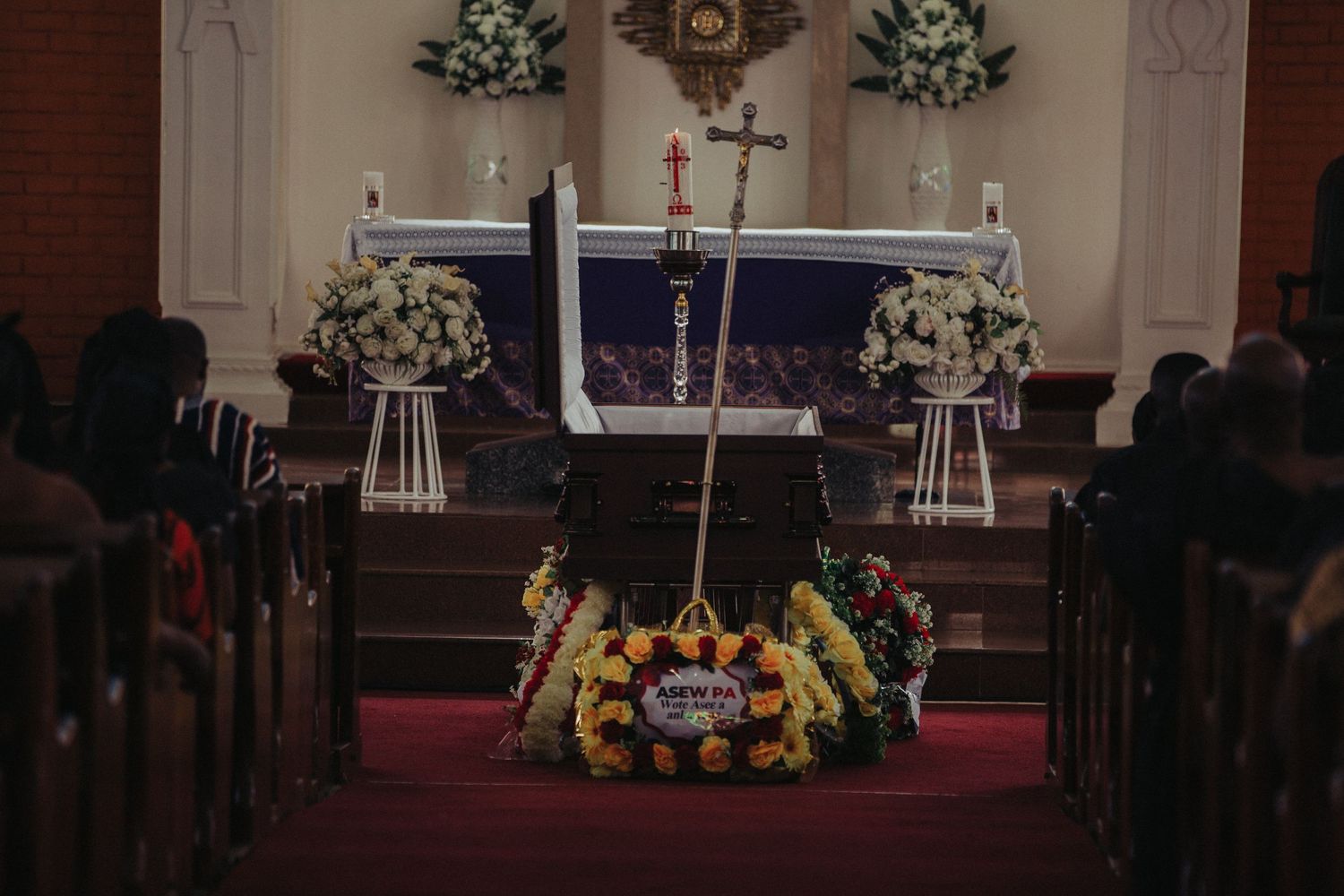 View of a casket at a church altar surrounded by white floral wreaths and a memorial cross.