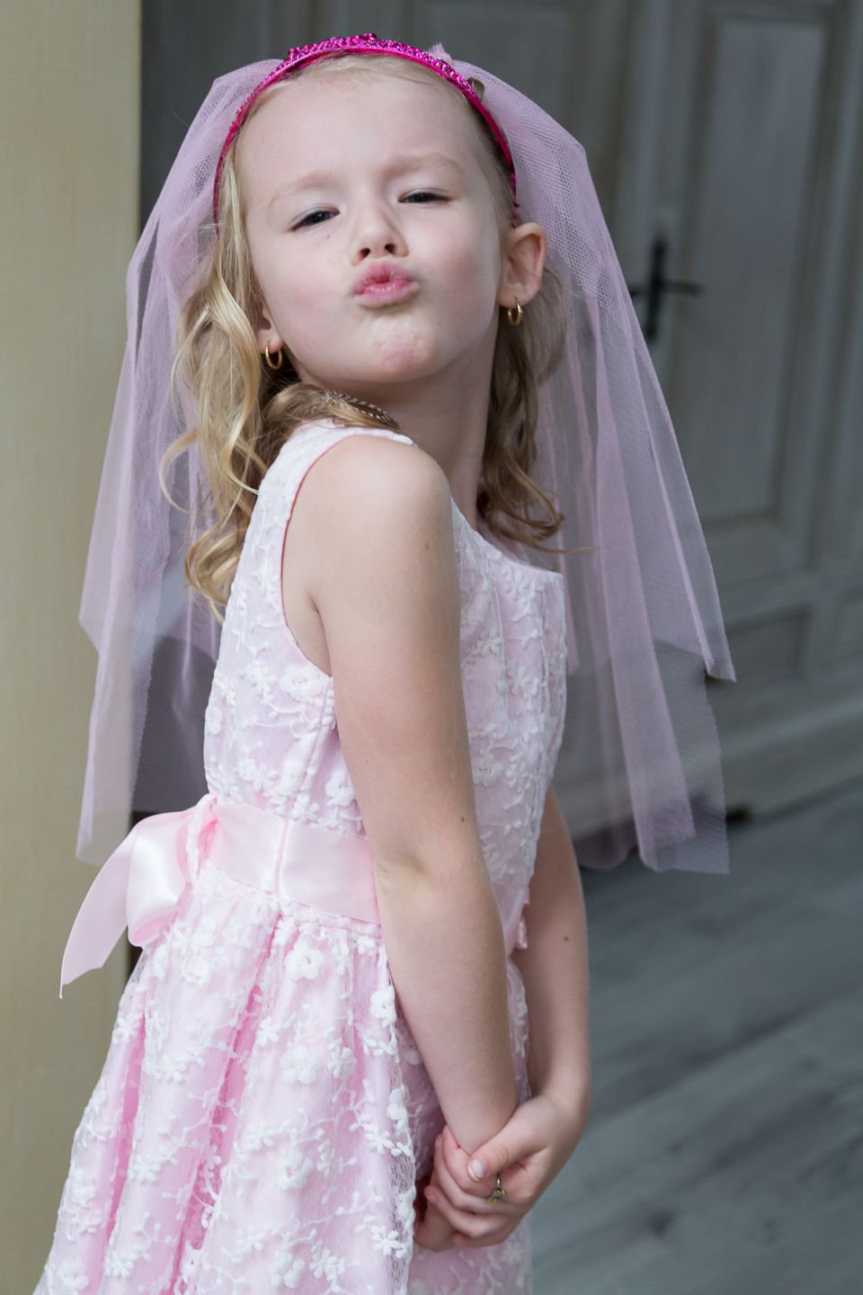 Young girl in white dress and pink veil making playful expression.