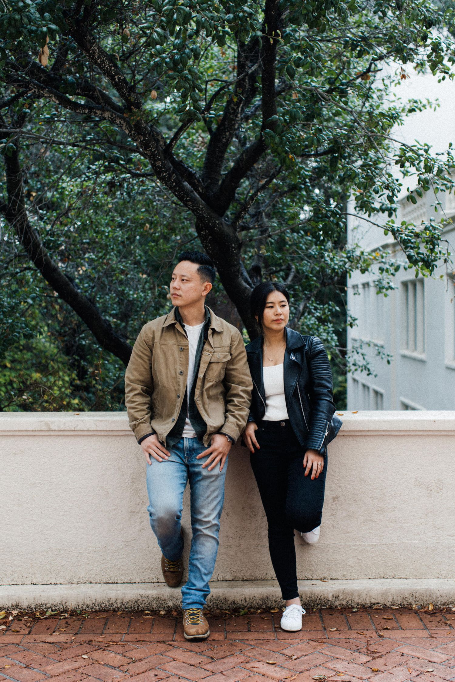 Casual couple in contrasting jackets posing against a brick wall under green trees.