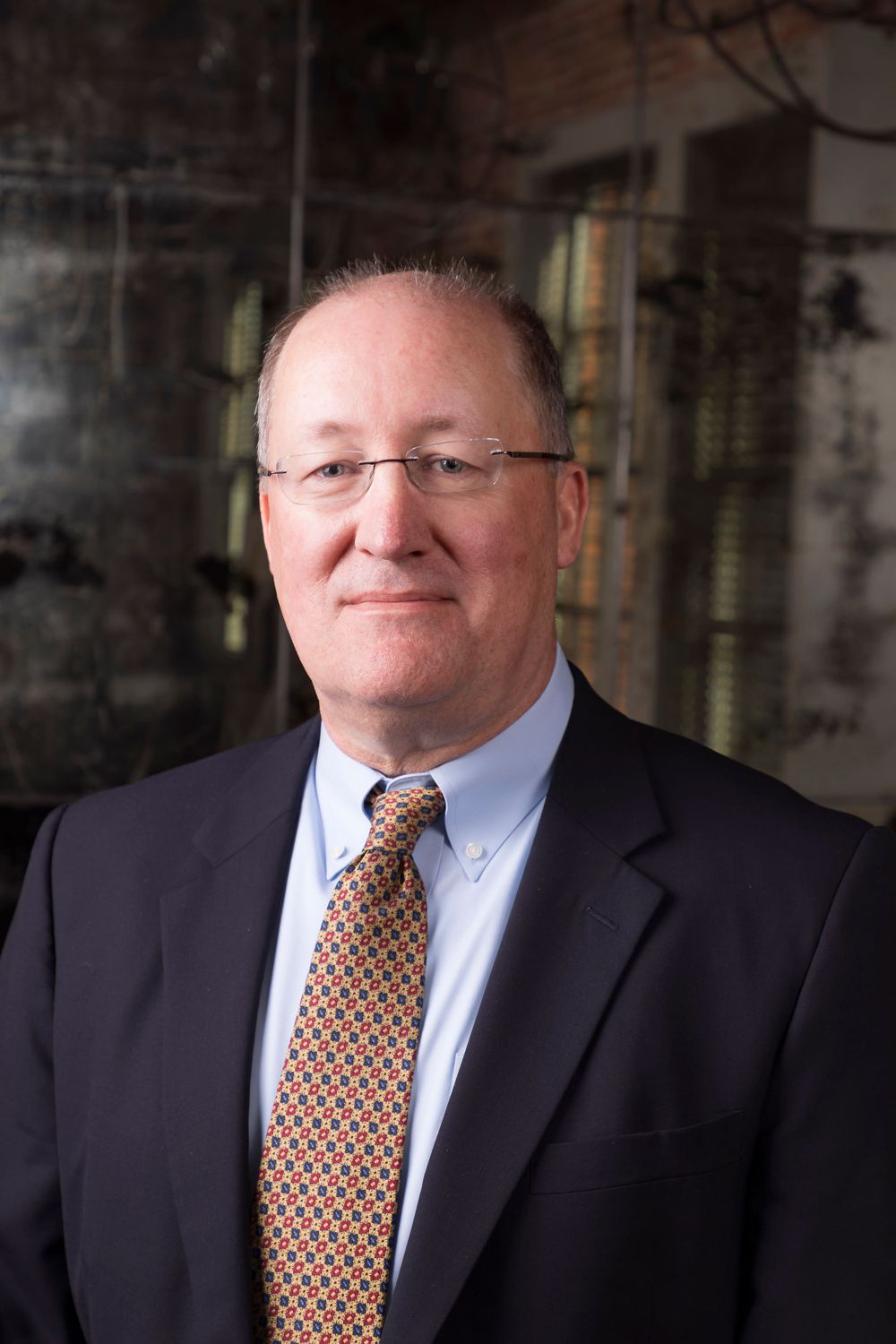 Business portrait of a person wearing navy suit and patterned tie against dark interior background.