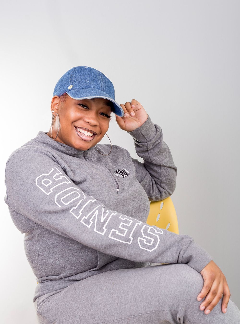 Person in gray Eagles sweatshirt and blue baseball cap posing playfully against light background.