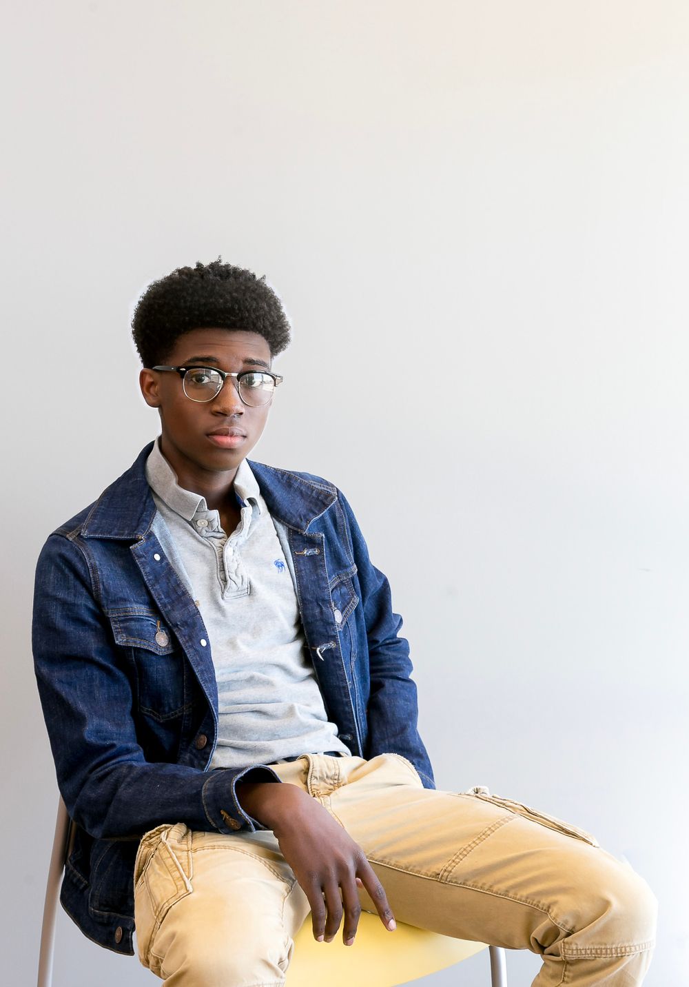 Stylish portrait of someone wearing a denim jacket, glasses and khaki pants against a light background.