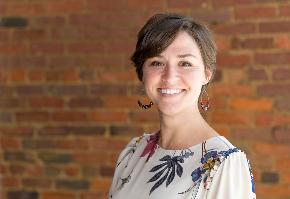 Professional portrait against a brick wall of someone wearing a floral patterned blouse and earrings.