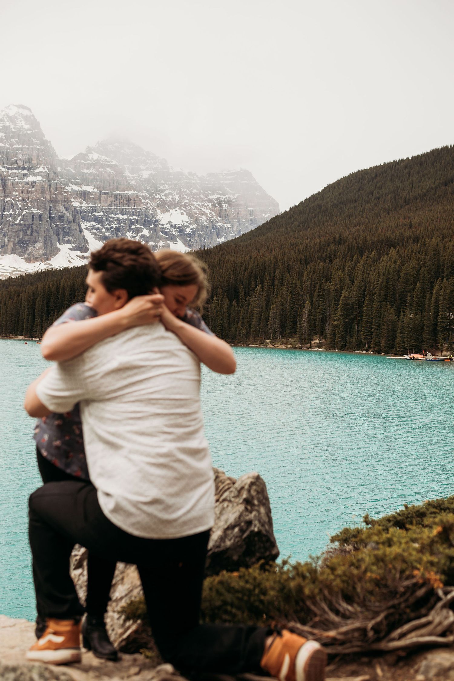 An intimate embrace captured at a scenic mountain lake overlook with evergreen forests and snowy peaks.
