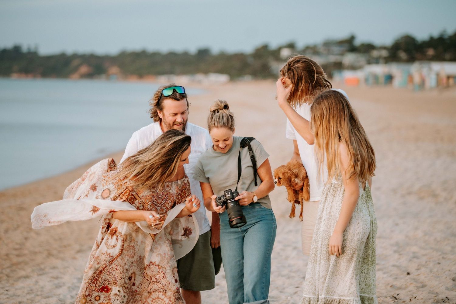 Three friends laugh and play with their small dog on a sunny beach at golden hour.